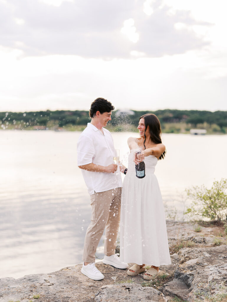 Lake Travis proposal photography featuring a couple popping champagne by the lakeside after saying yes.