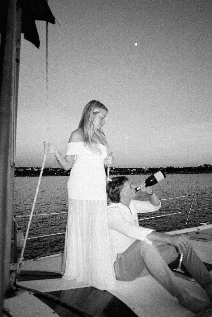 Couple popping champagne during their Lake Travis sailboat engagement session, captured at sunset.