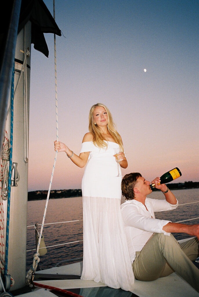Couple popping champagne during their Lake Travis sailboat engagement session, captured at sunset.
