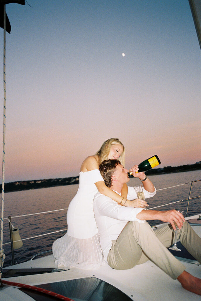 Couple popping champagne during their Lake Travis sailboat engagement session, captured at sunset.