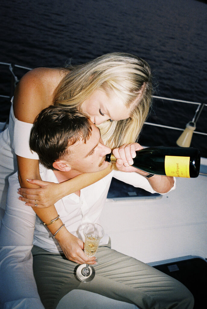 Couple popping champagne during their Lake Travis sailboat engagement session, captured at sunset.