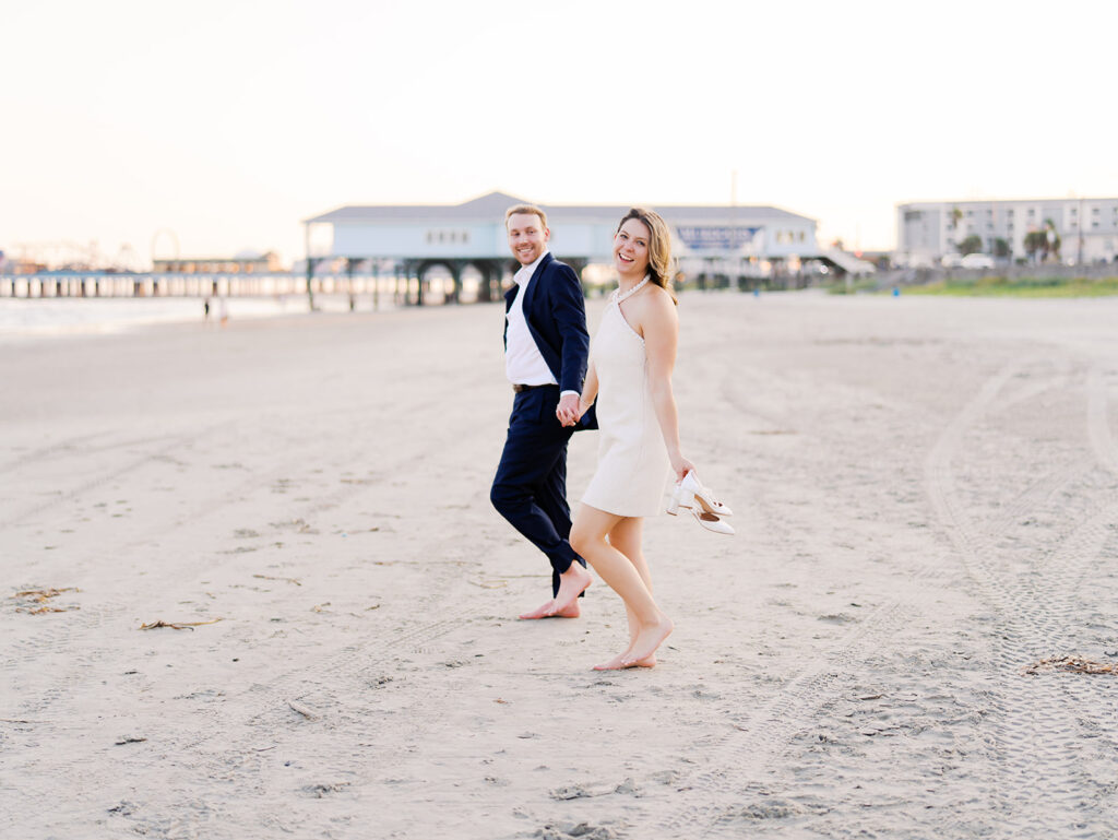 Emma and Luke walking hand in hand along the beach in front of the Grand Galvez at sunset with soft pastel tones on film