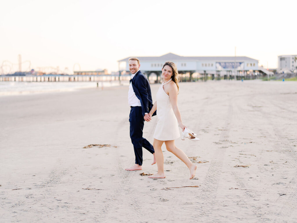 Emma and Luke walking hand in hand along the beach in front of the Grand Galvez at sunset with soft pastel tones on film