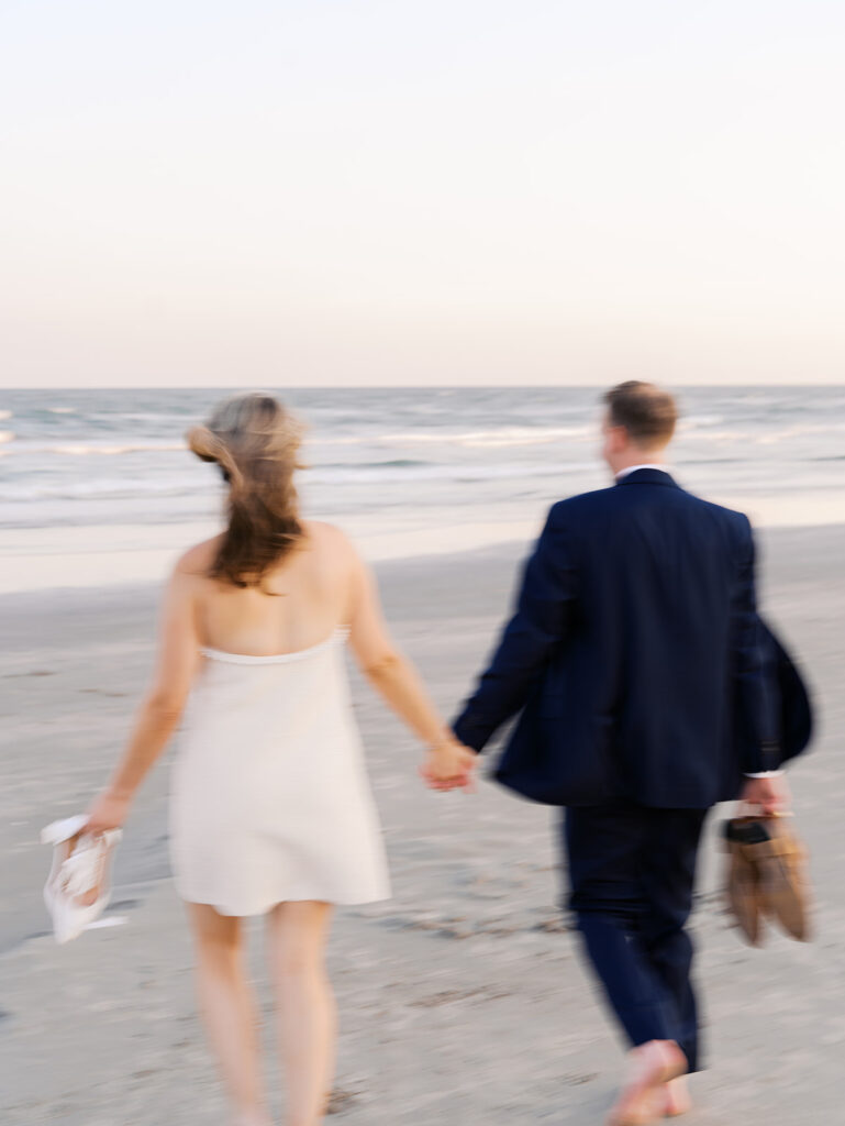 Emma and Luke walking hand in hand along the beach in front of the Grand Galvez at sunset with soft pastel tones on film