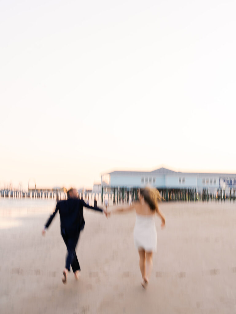 Emma and Luke walking hand in hand along the beach in front of the Grand Galvez at sunset with soft pastel tones on film