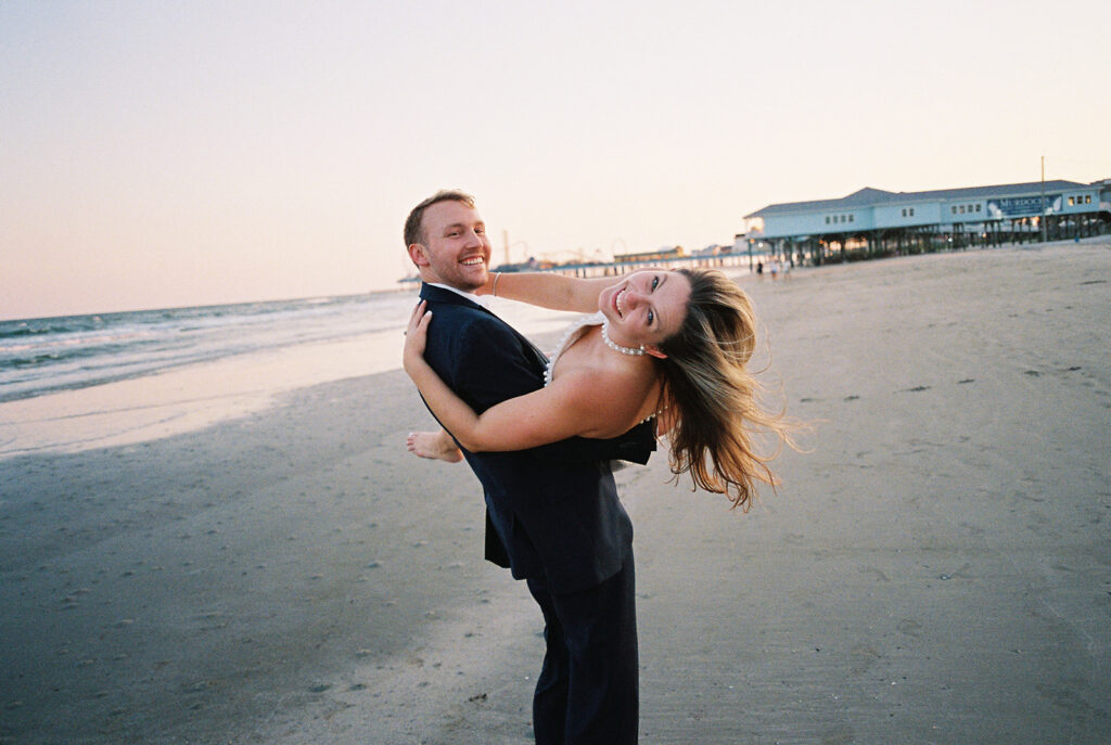 Emma and Luke walking hand in hand along the beach in front of the Grand Galvez at sunset with soft pastel tones on film