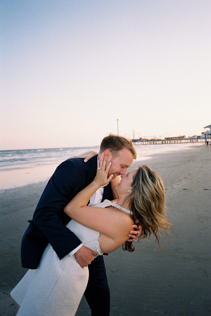 Emma and Luke walking hand in hand along the beach in front of the Grand Galvez at sunset with soft pastel tones on film