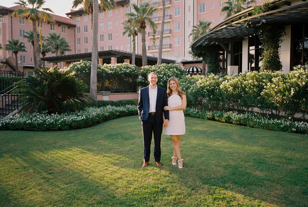 Emma and Luke smiling together in the Grand Galvez garden under tall palm trees captured on film with soft, natural light and timeless color.